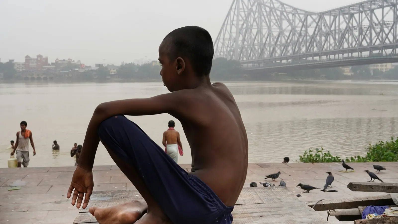 A boy sitting by the river with the Howrah Bridge behind