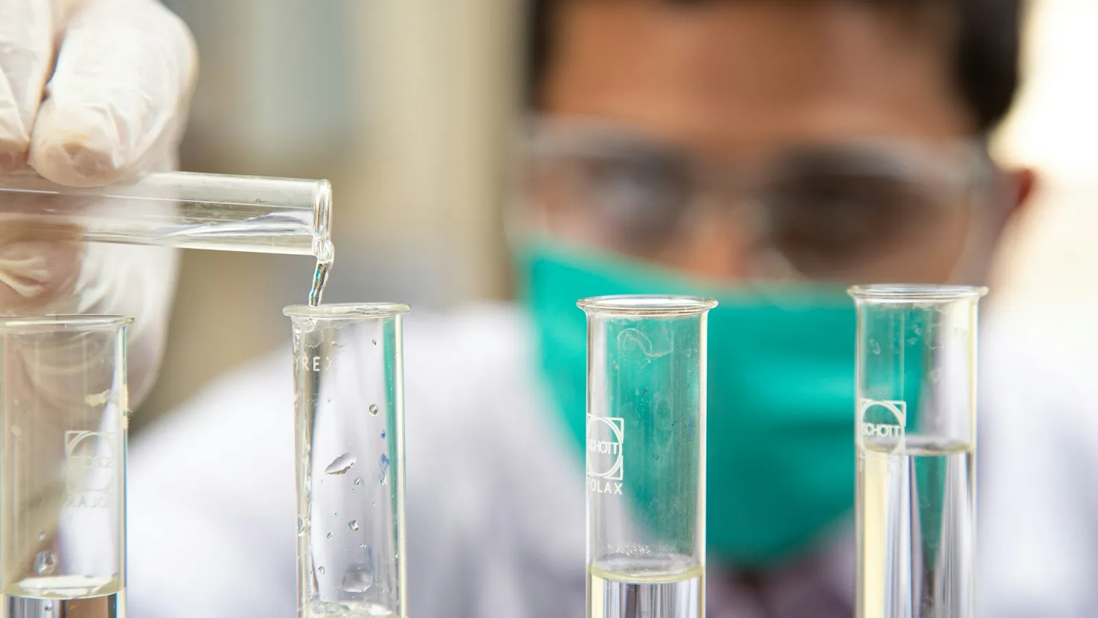 A chemist in goggles and a mask pouring liquid into test tubes