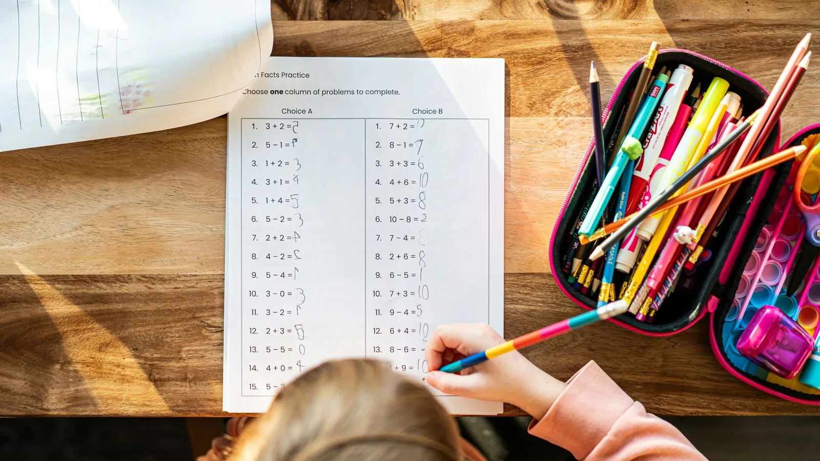 A child working on a maths worksheet beside a case of coloured pencils