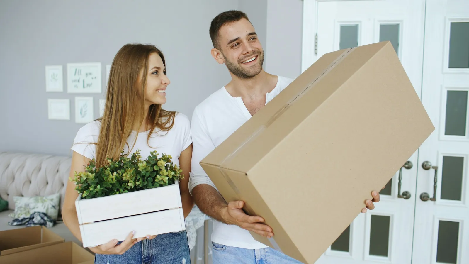 A couple carrying a cardboard box and a plant into a new home