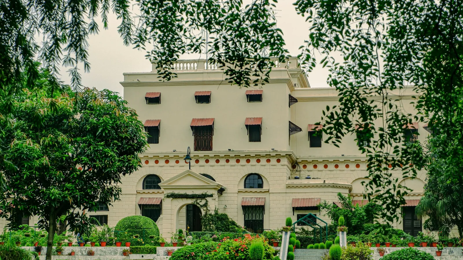 A cream colonial school building behind trees and a garden