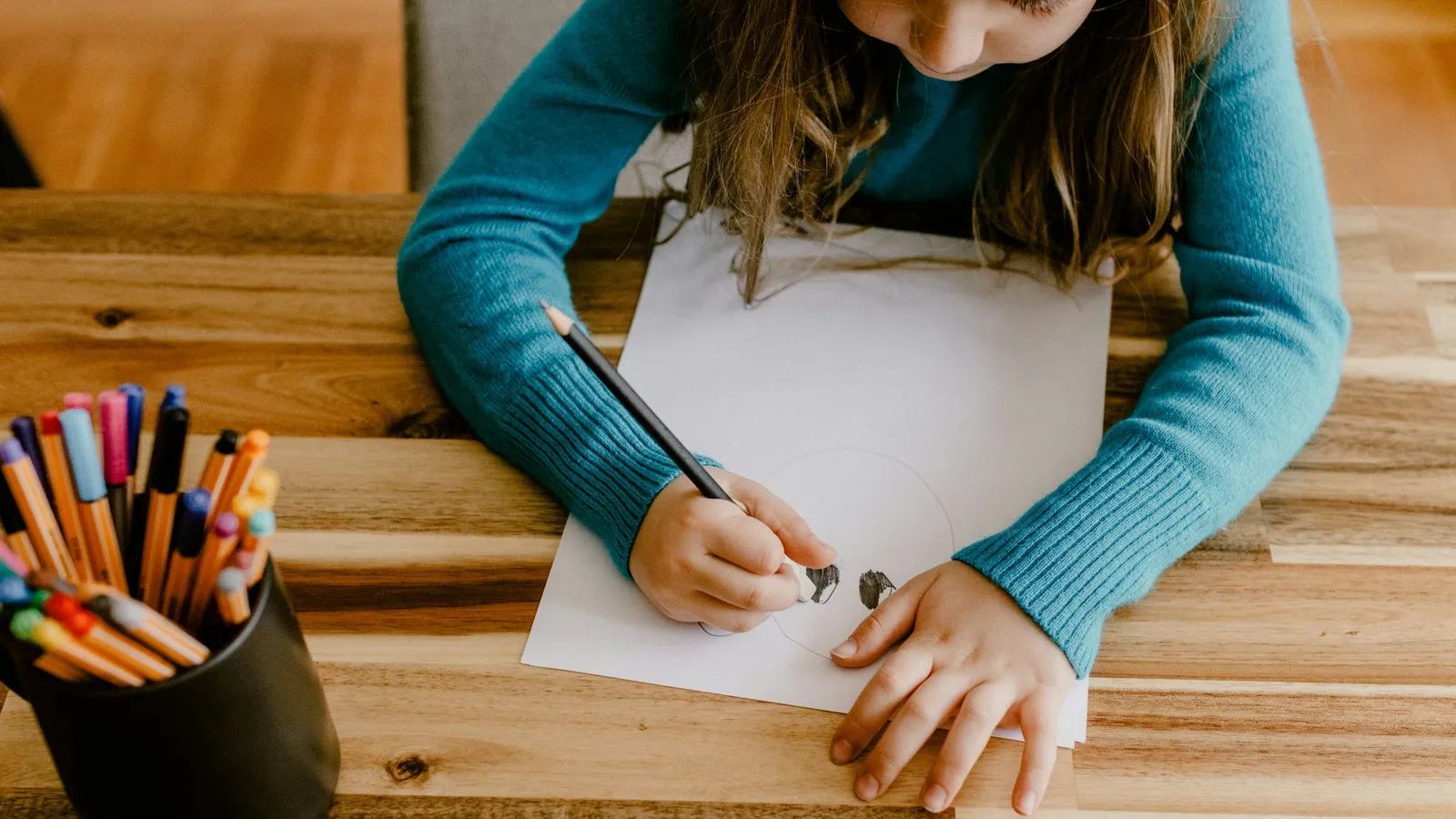 A girl drawing at a table with a pot of coloured pencils