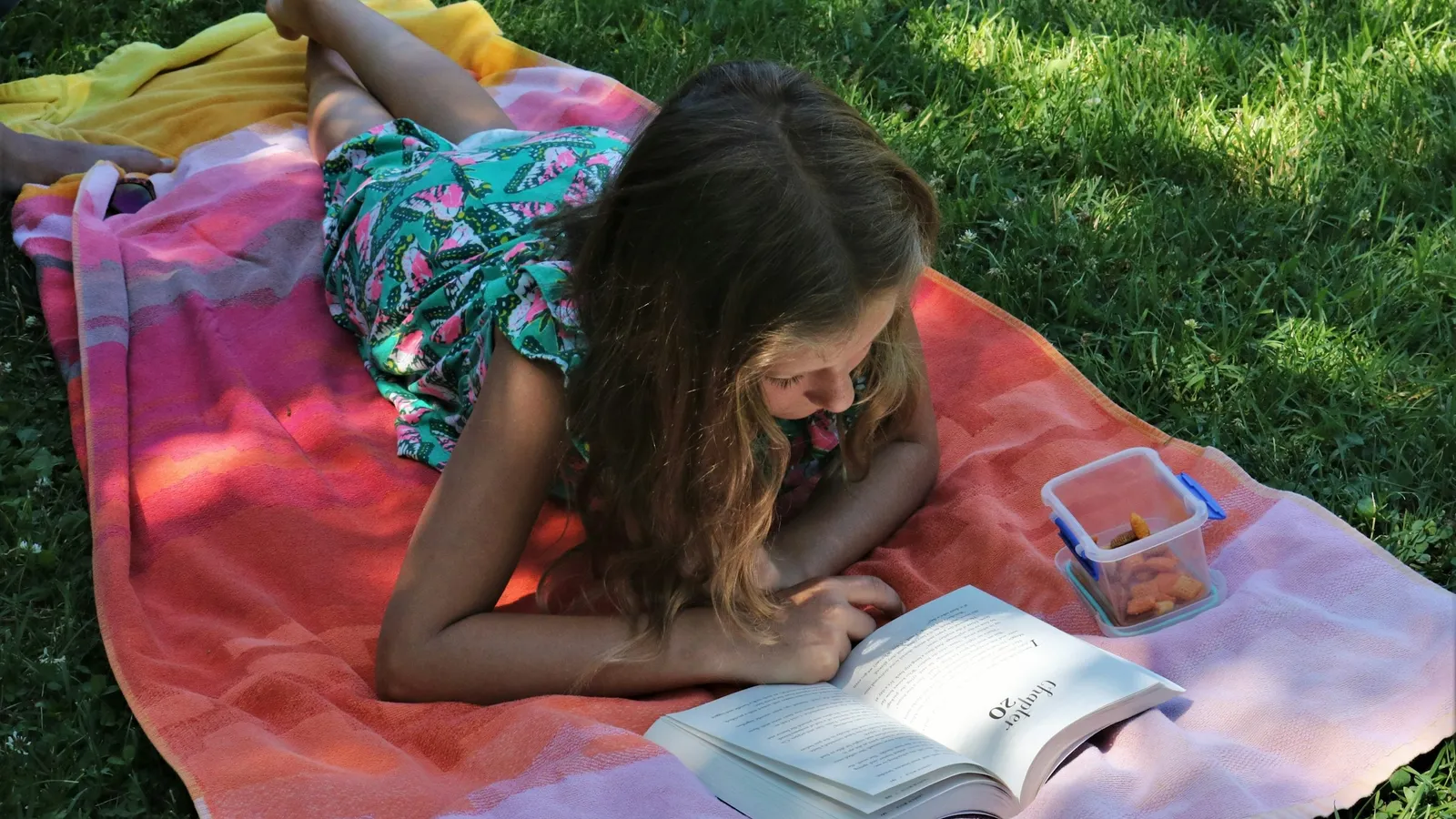A girl lying on a blanket on the grass, reading a book