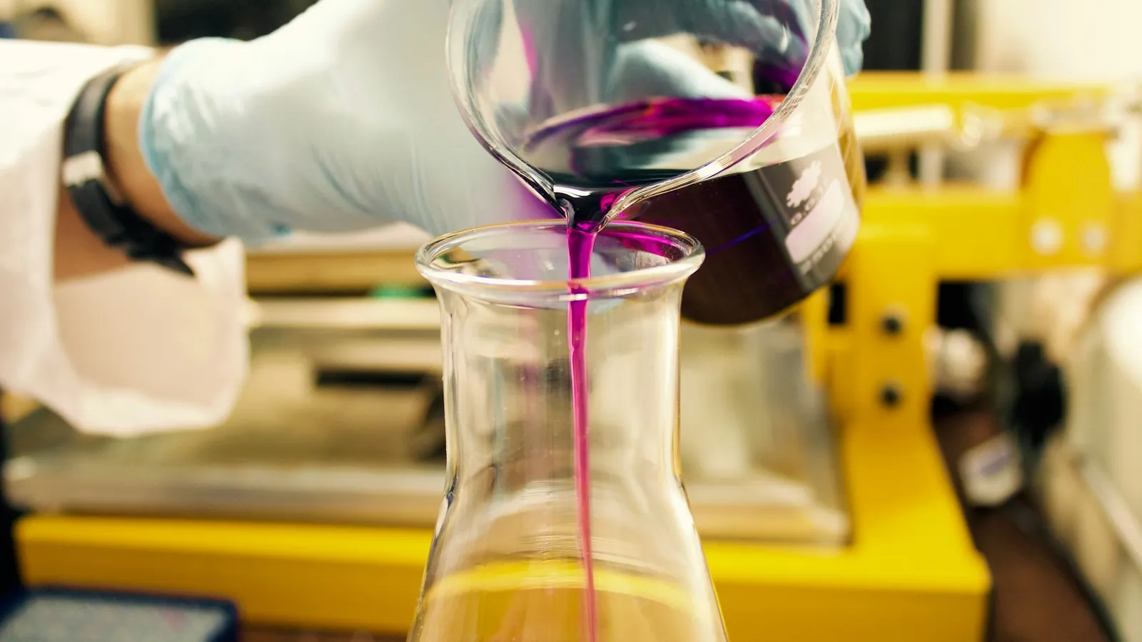 A gloved hand pouring purple liquid into a glass flask