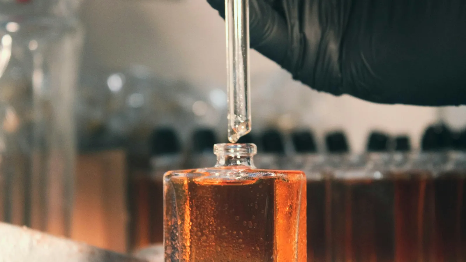 A gloved hand using a dropper over a glass of amber liquid