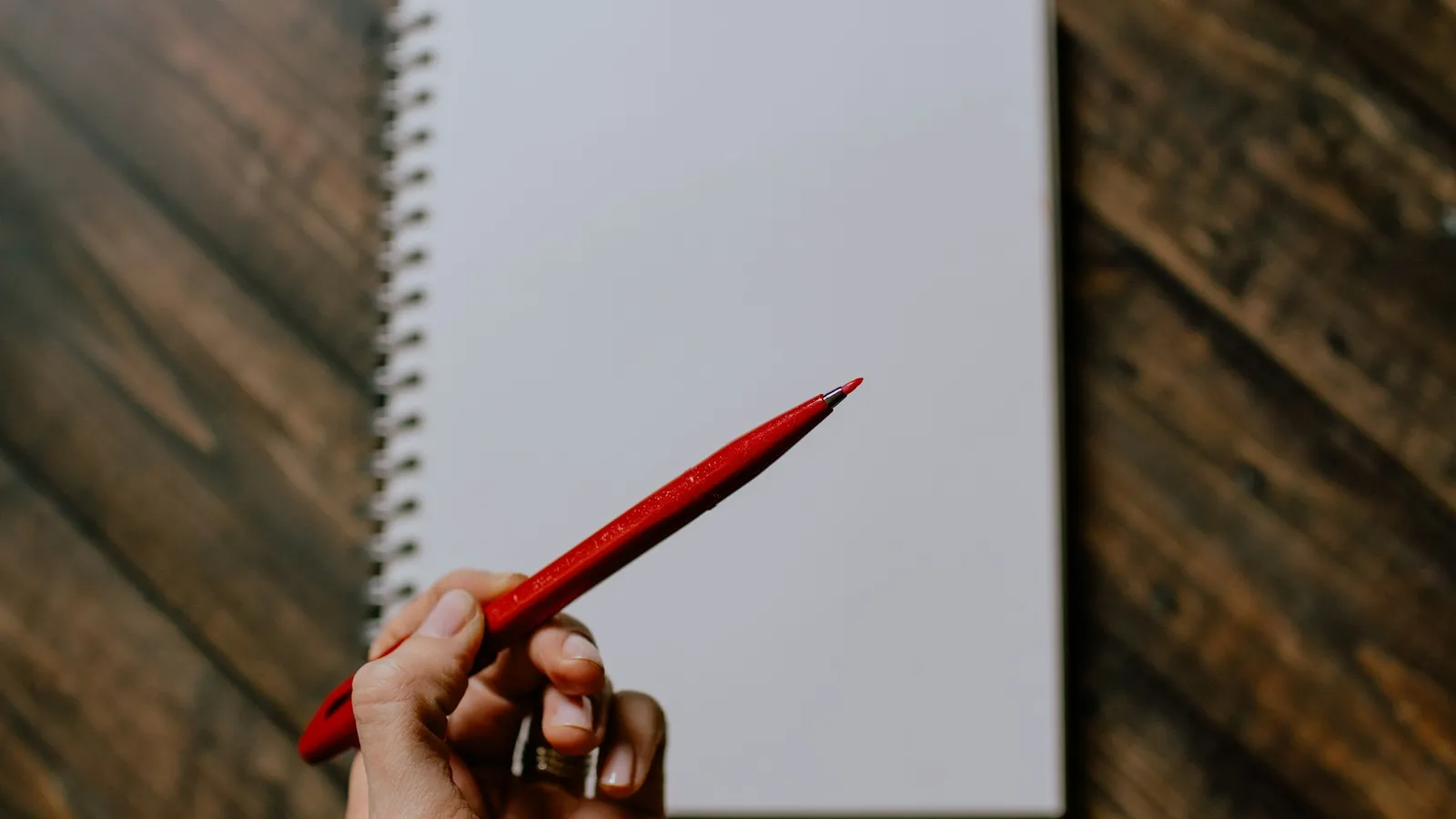 A hand holding a red pen over a blank spiral notebook