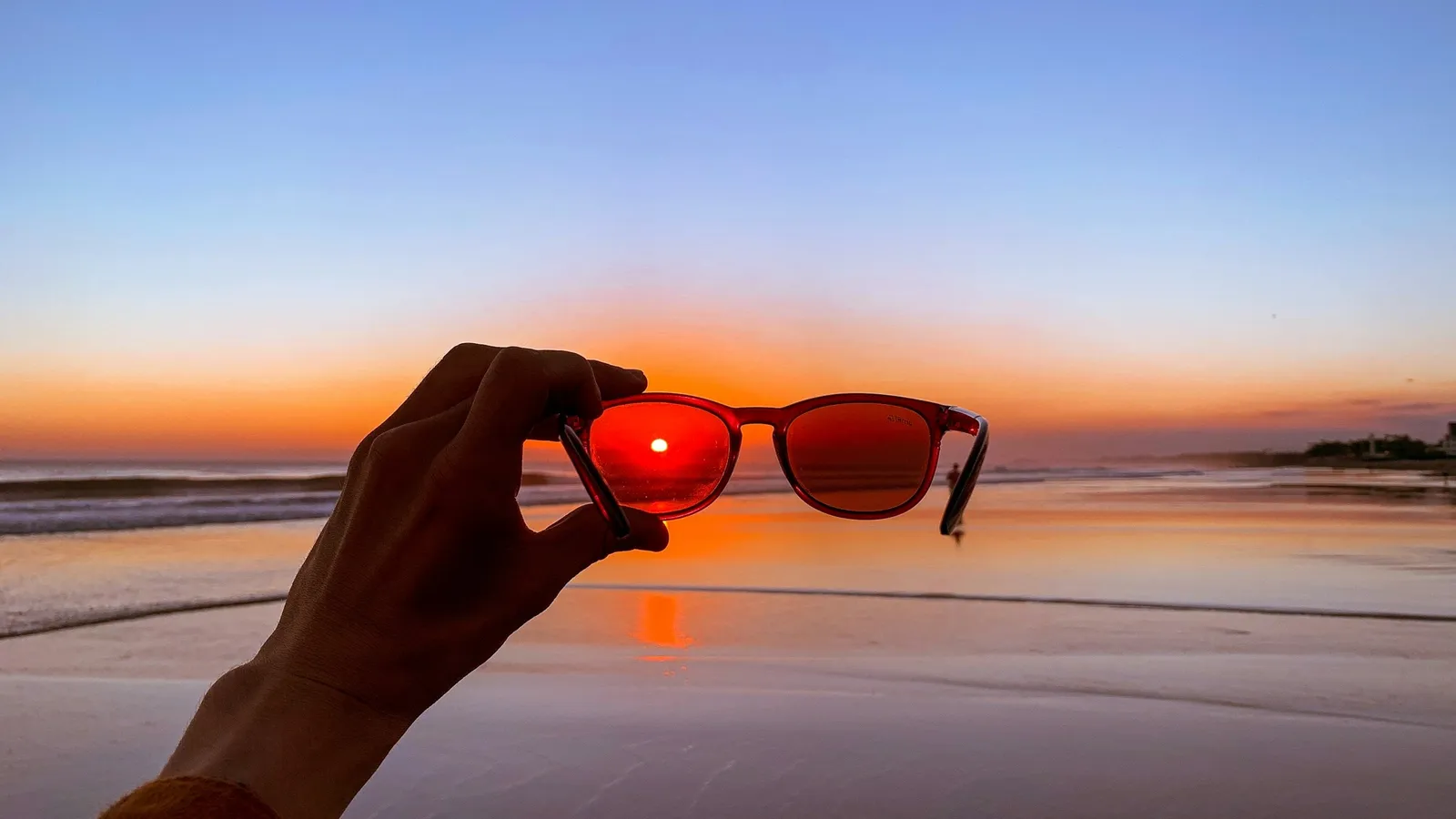A hand holding up sunglasses against an orange sunset over the sea