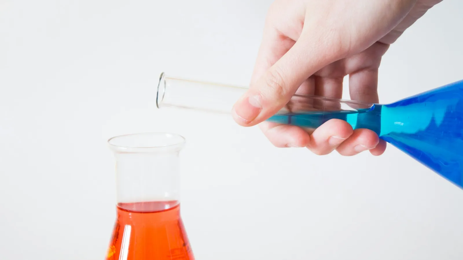 A hand pouring blue liquid from a test tube into a flask of orange liquid