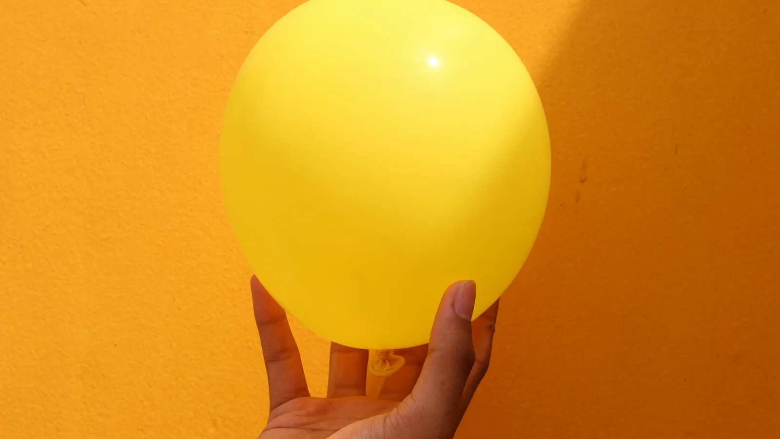 A hand pressing on a yellow balloon against an orange wall