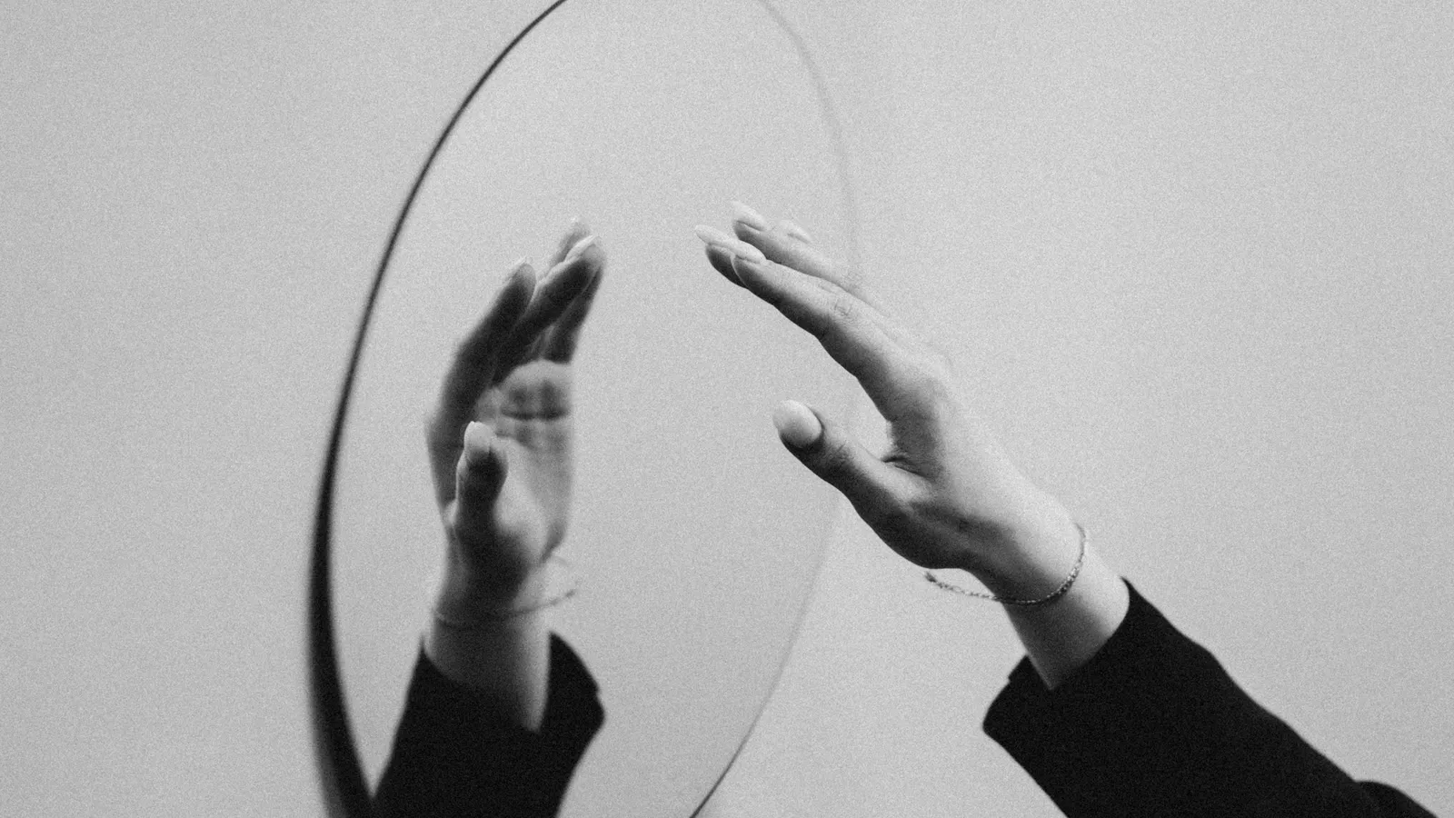 A hand reaching towards its reflection in a round mirror, in black and white