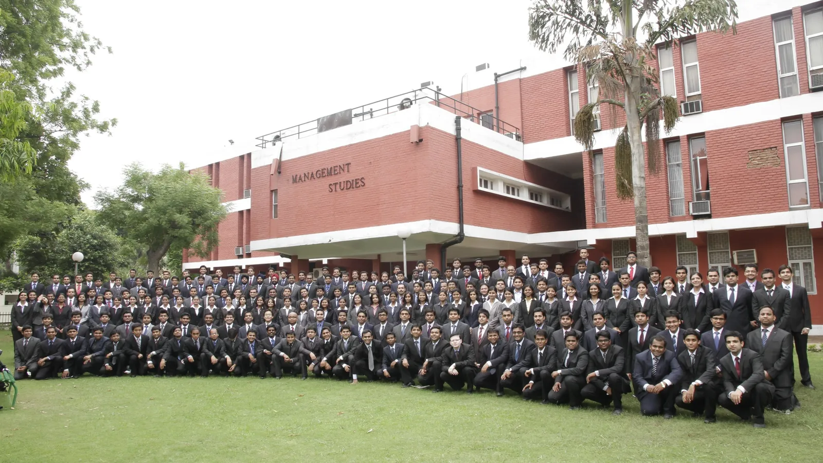 A large new batch of students in suits posing on a lawn outside a red-brick college building