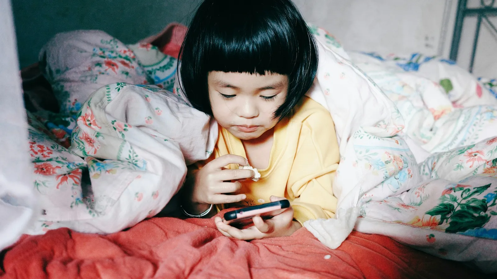 A little girl lying in bed looking at a phone