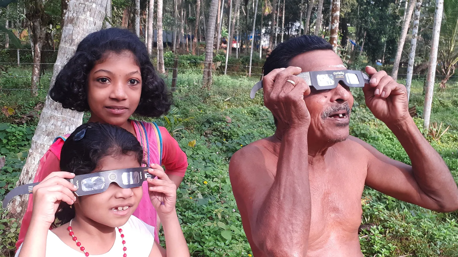 A man and a young girl watch a solar eclipse through eclipse glasses while an older girl stands beside them