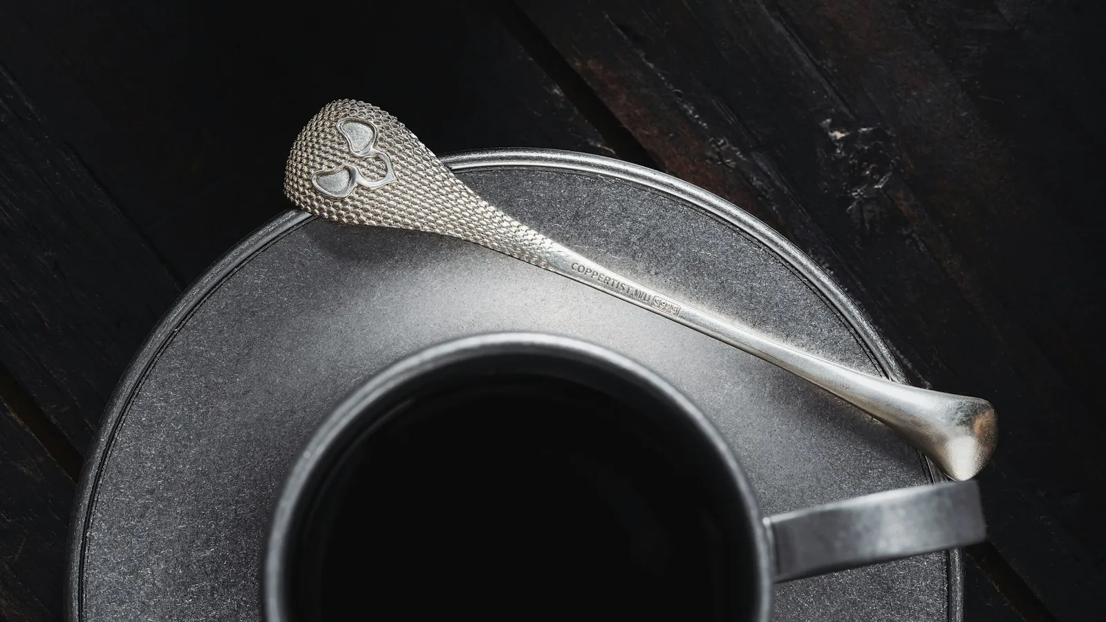 A metal spoon resting on the saucer of a cup of black coffee