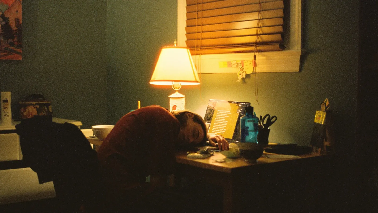 A person slumped at a desk under a lamp in a dim room