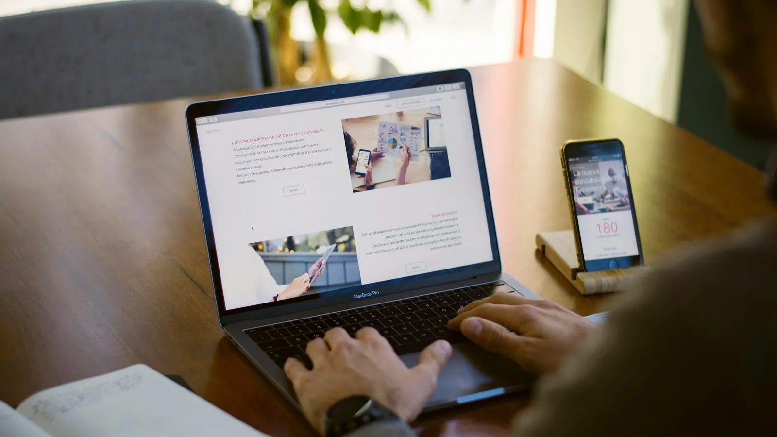 A person typing on a laptop with a smartphone propped up beside it