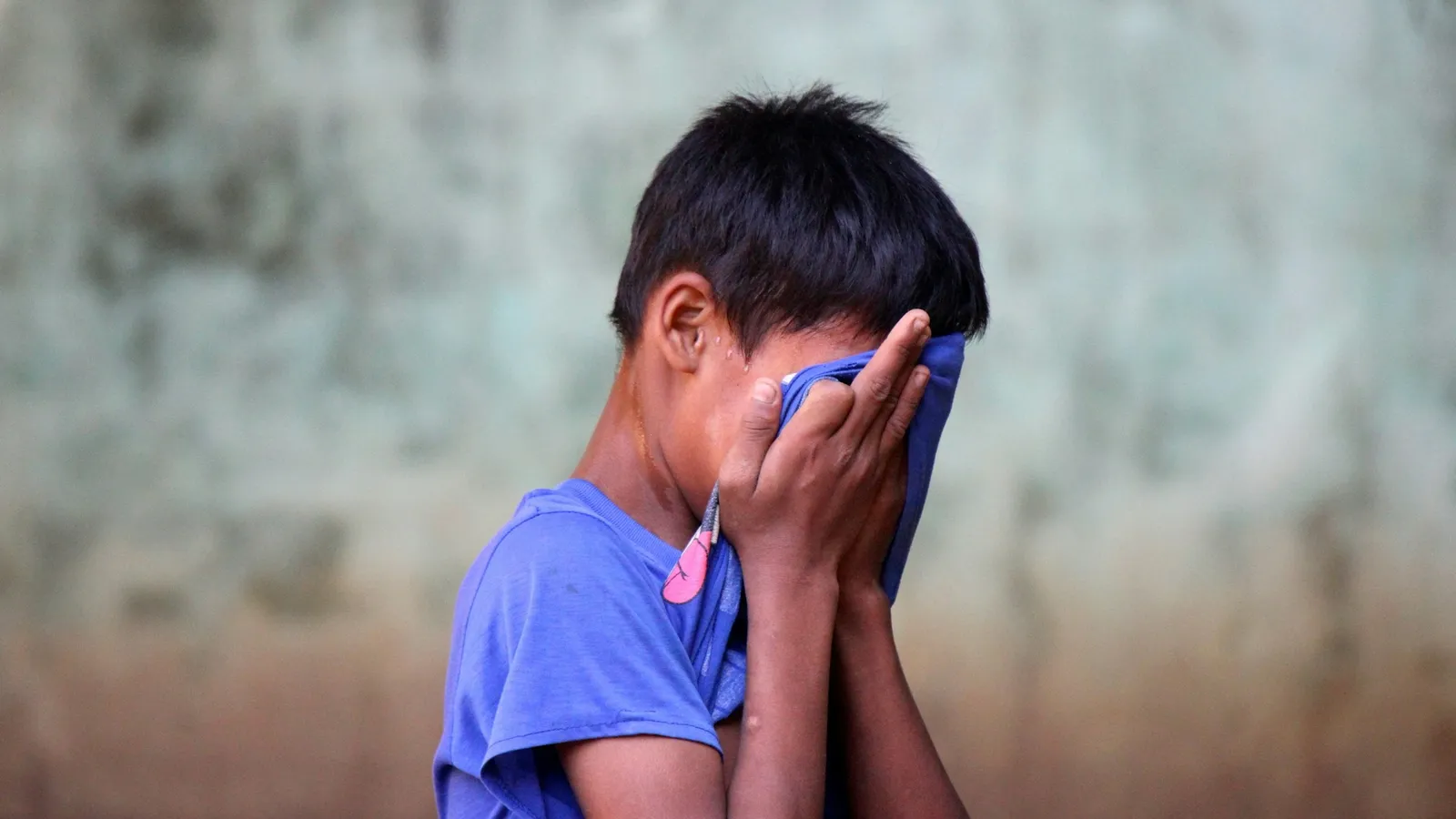 A shy boy in a blue shirt covering his face with his hands