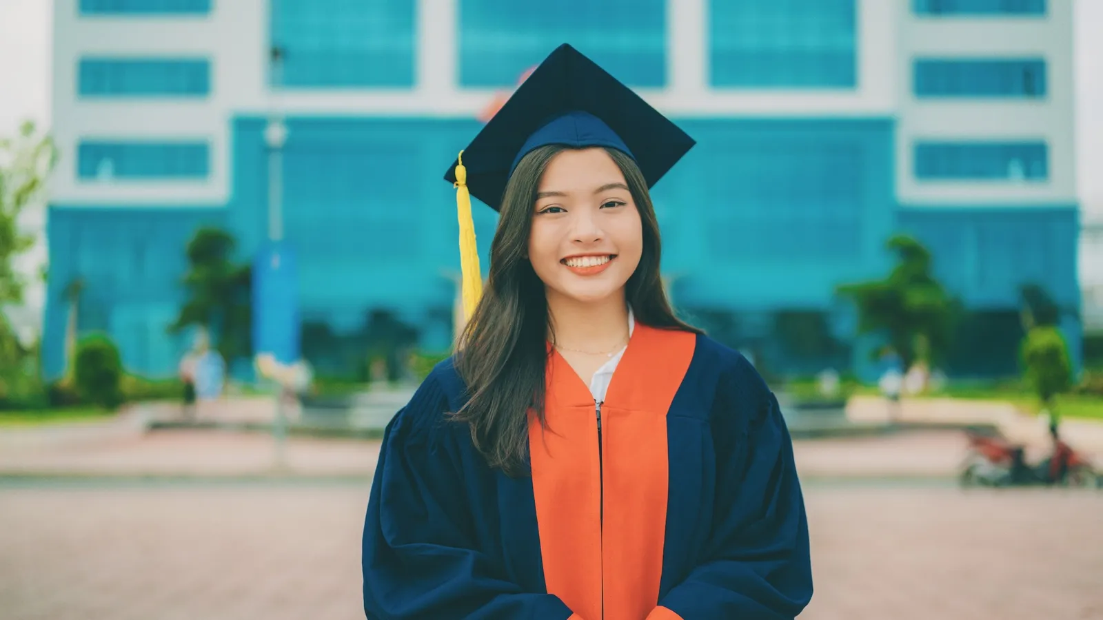 A smiling graduate in cap and gown outside a college building