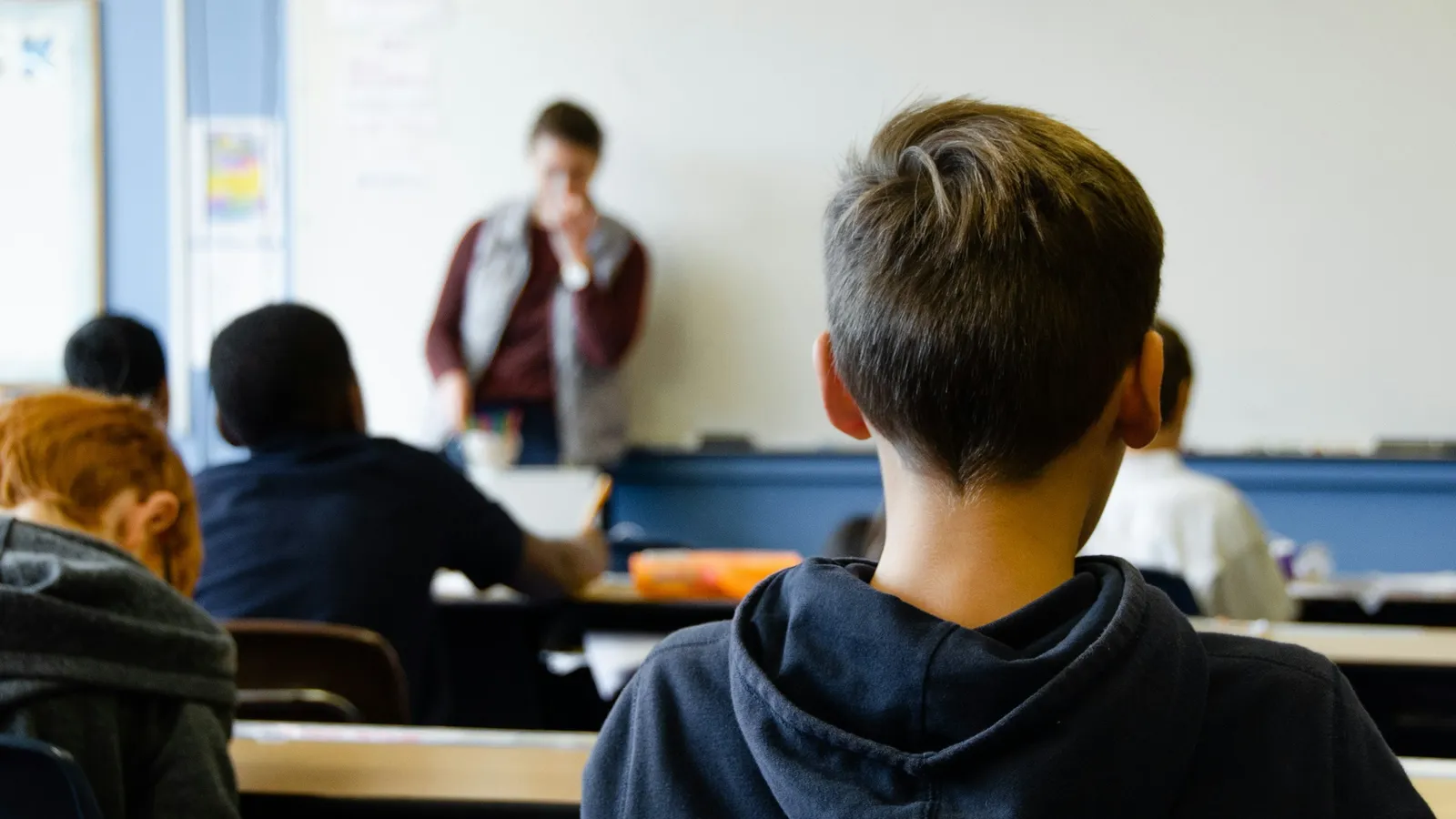 A student in a hoodie watching a teacher at the front of a classroom