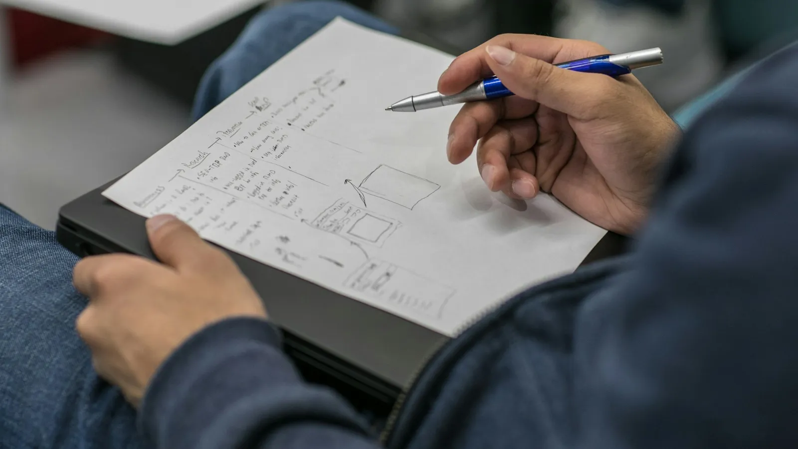 A student writing notes and a diagram on paper resting on their lap