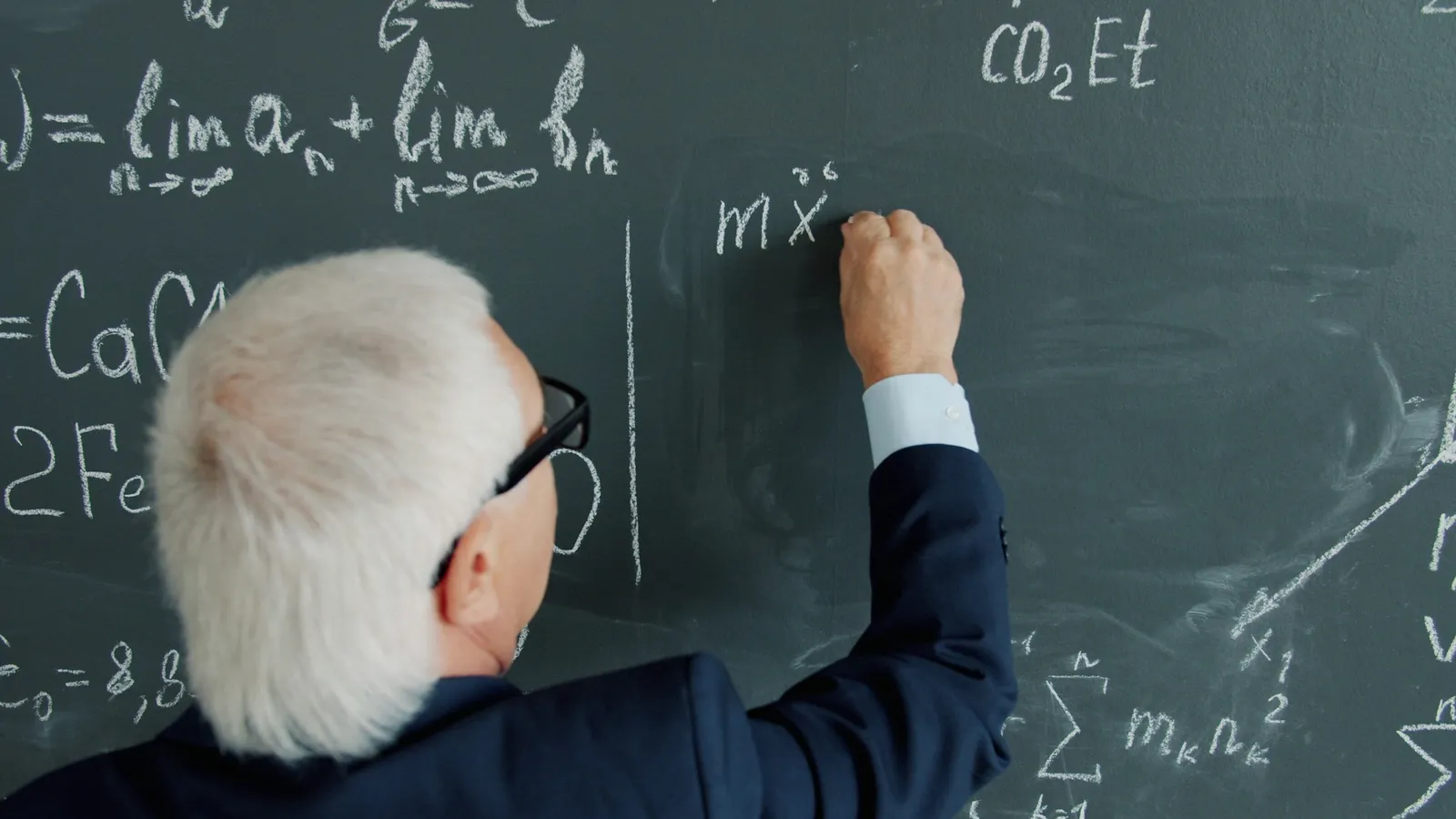A teacher writing equations on a green chalkboard