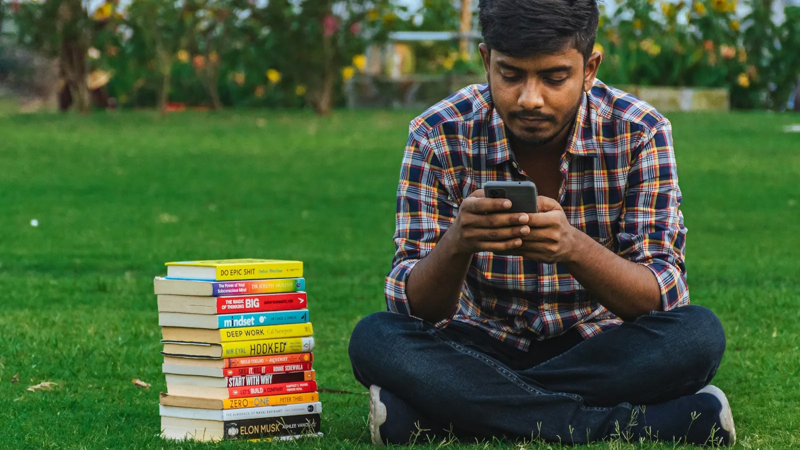 A teenager sitting on grass beside a stack of books, looking at a phone