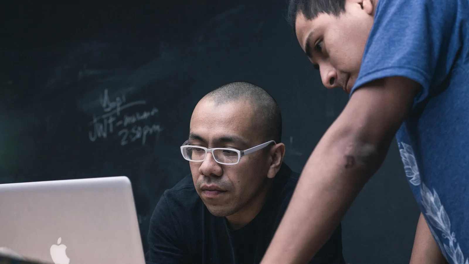 A tutor at a laptop explaining a problem to a student beside a chalkboard