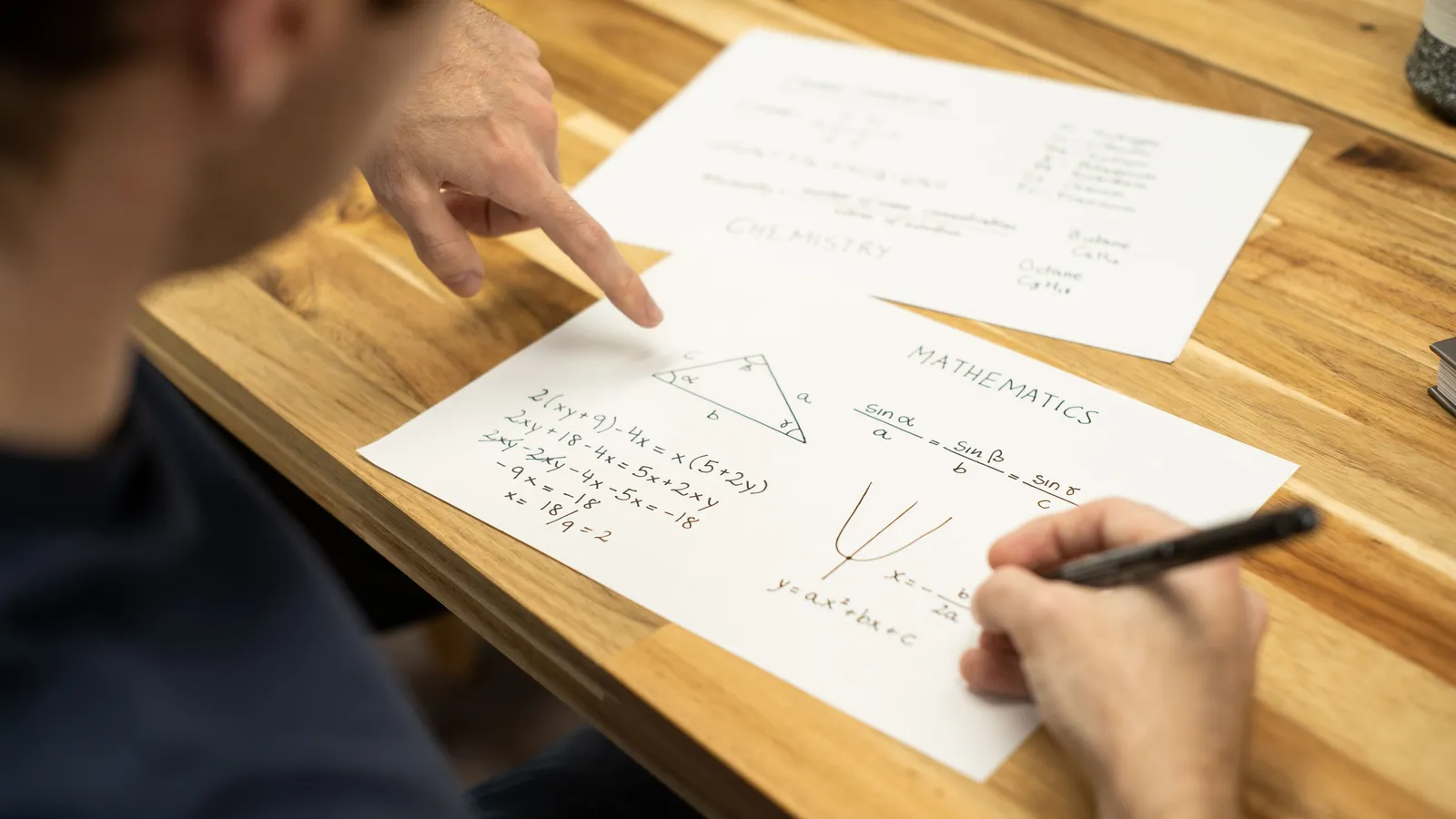 A tutor's finger points at a student's handwritten maths working while the student writes