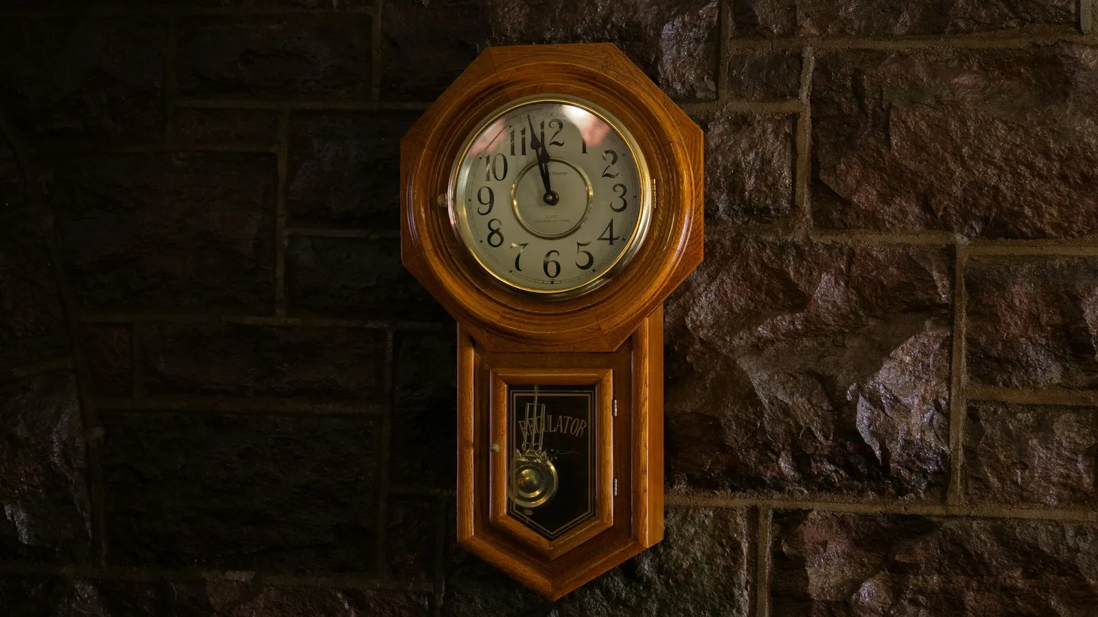 A wooden pendulum wall clock hanging on a stone wall