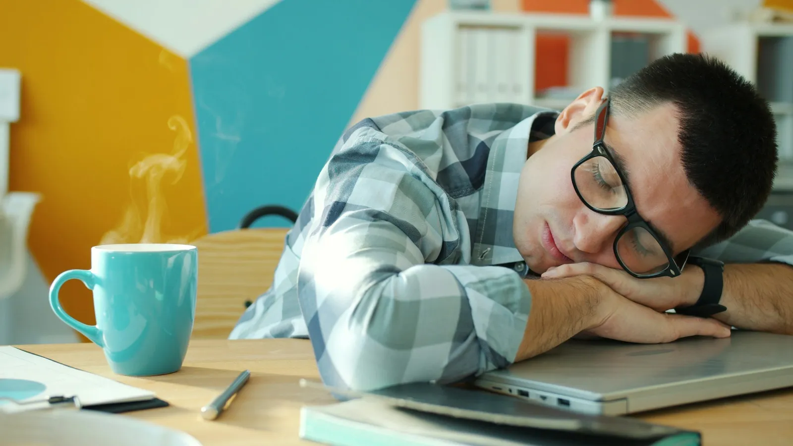 A young man asleep with his head on a desk beside a laptop and a mug
