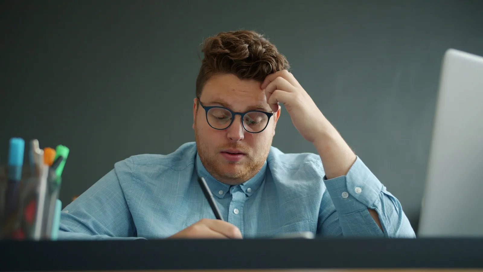 A young man in glasses holding his head while working at a desk