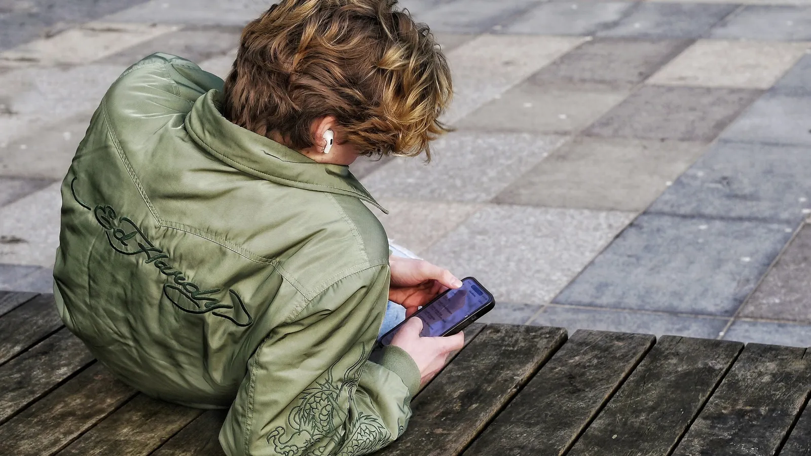A young person in a green jacket sitting alone looking at a phone