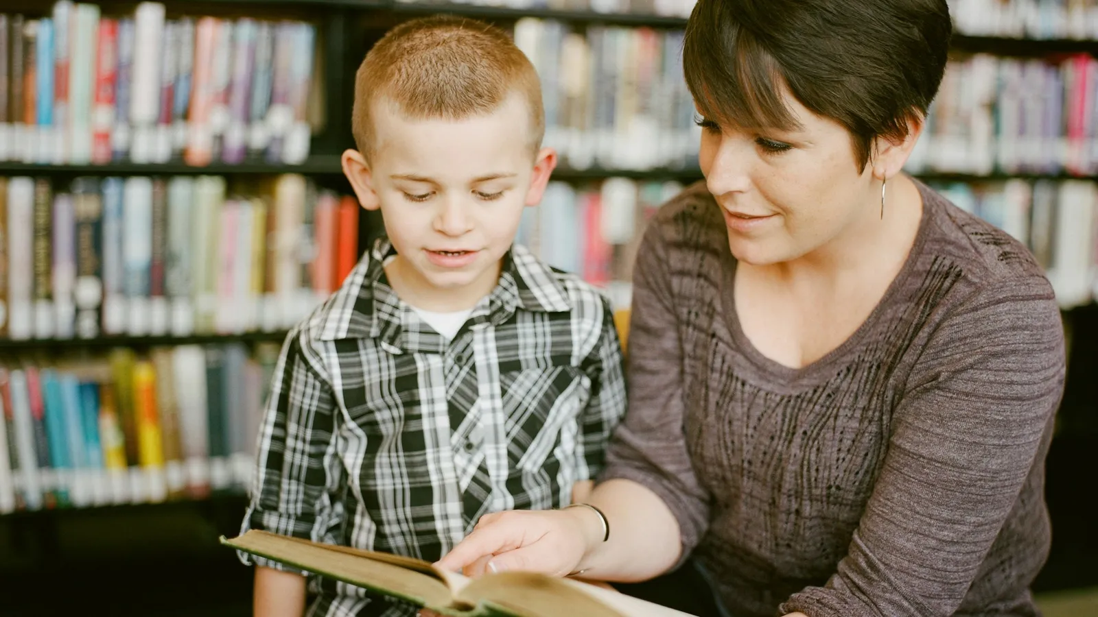 Adult helping a boy read a book in a library