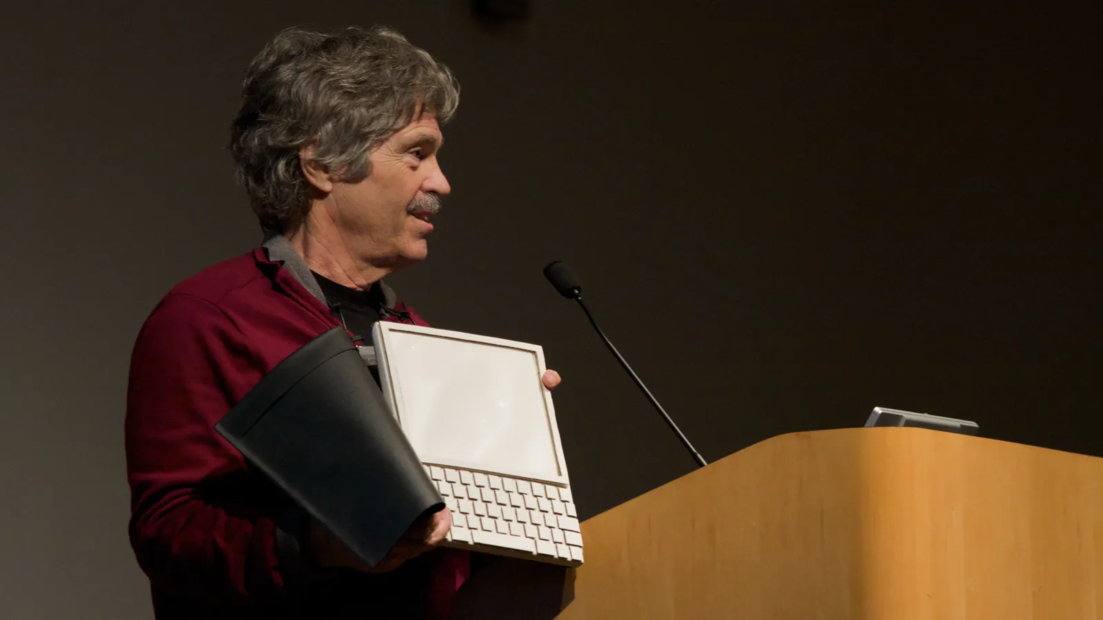 Alan Kay beside a lectern holding a white tablet-and-keyboard Dynabook model