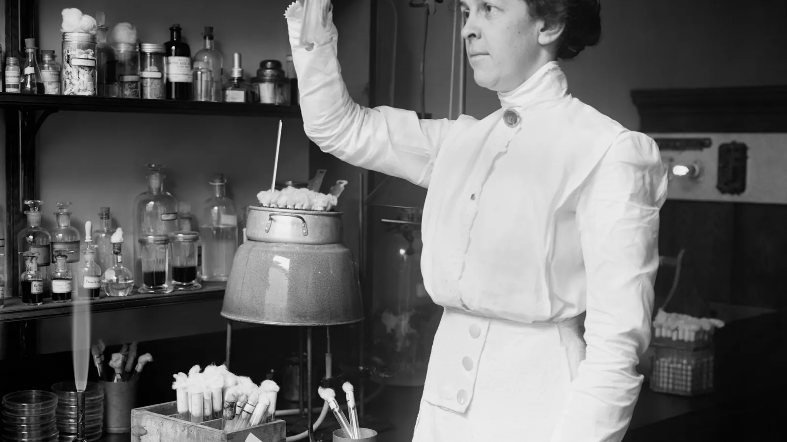 Alice Evans in a white dress holding up cotton-stoppered test tubes beside laboratory shelves of bottles