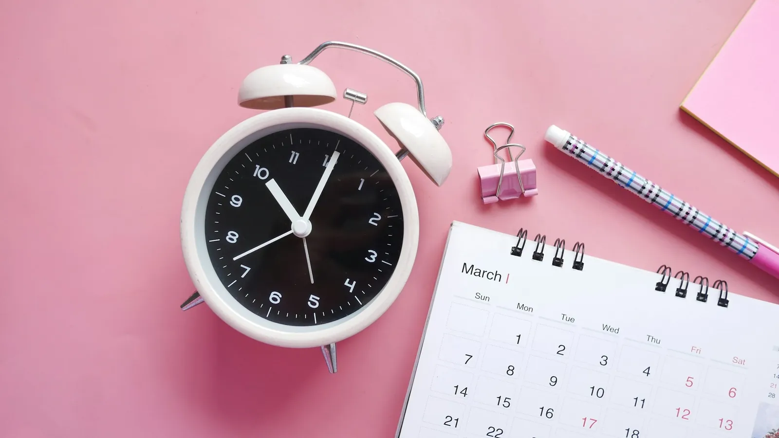 An alarm clock beside a calendar on a pink desk