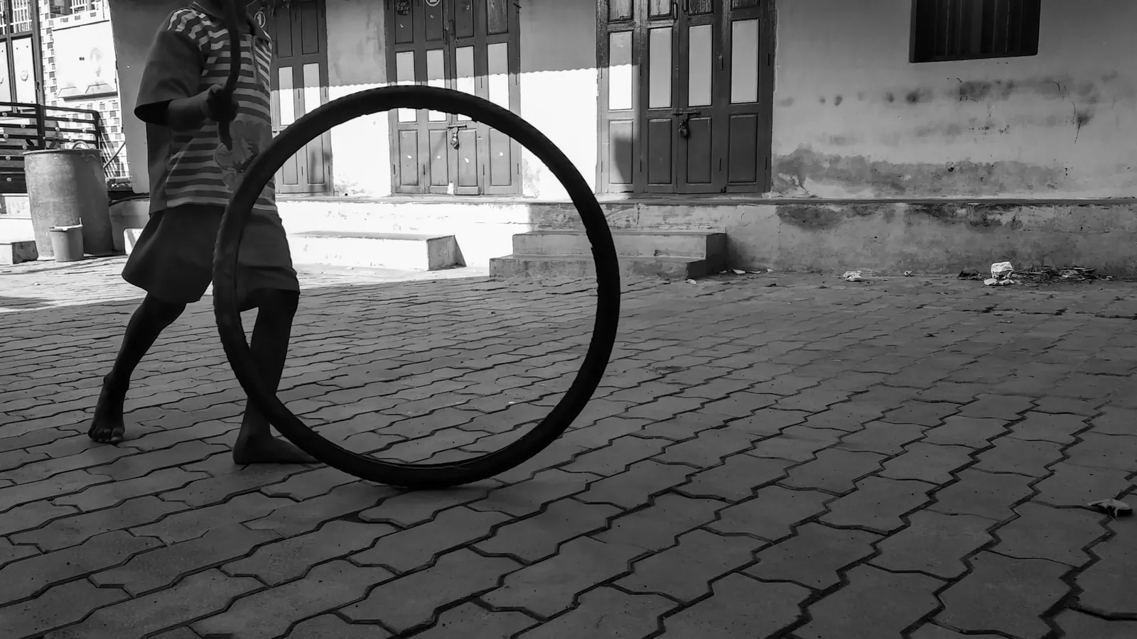 Black-and-white photo of a barefoot boy rolling a bicycle tyre along a street