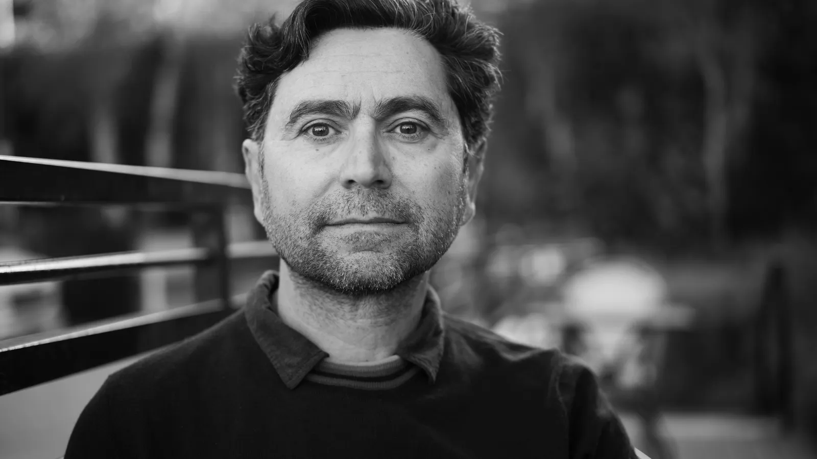 Black-and-white portrait of Ardem Patapoutian seated outdoors beside a metal railing