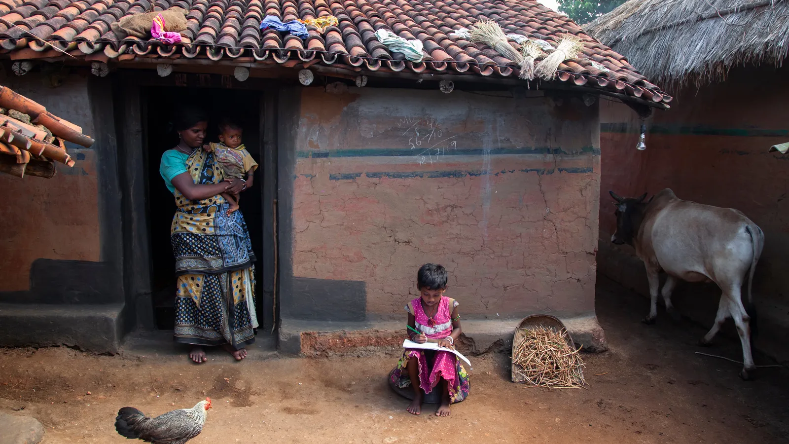 Child writing in a notebook outside a village home while a mother holds a baby