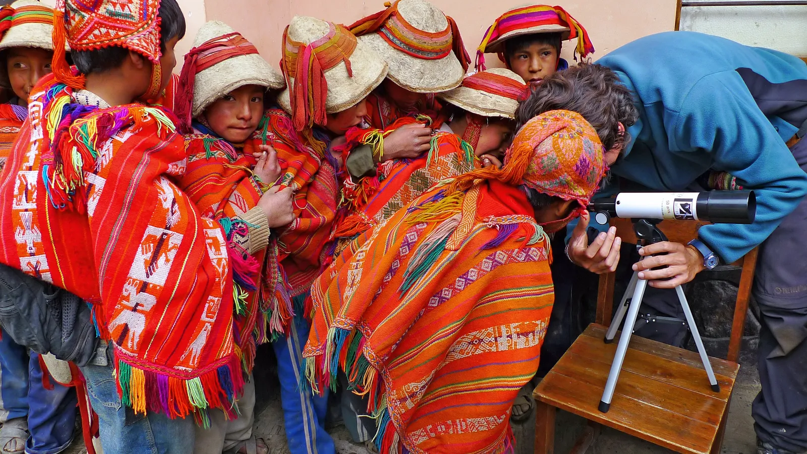 Children in colourful ponchos and hats crowd around a small telescope on a chair