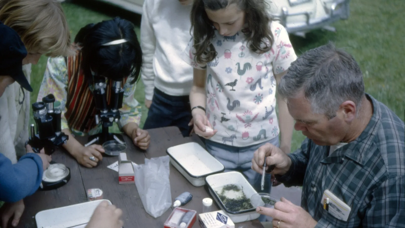 Children use microscopes while an adult examines pond samples with them at an outdoor table
