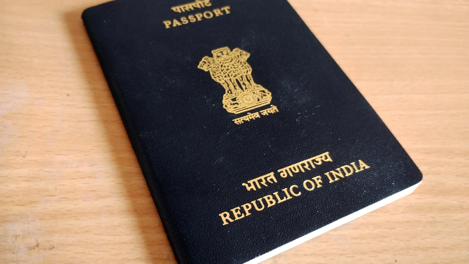 Closed Indian passport with a gold national emblem lying on a light wooden table