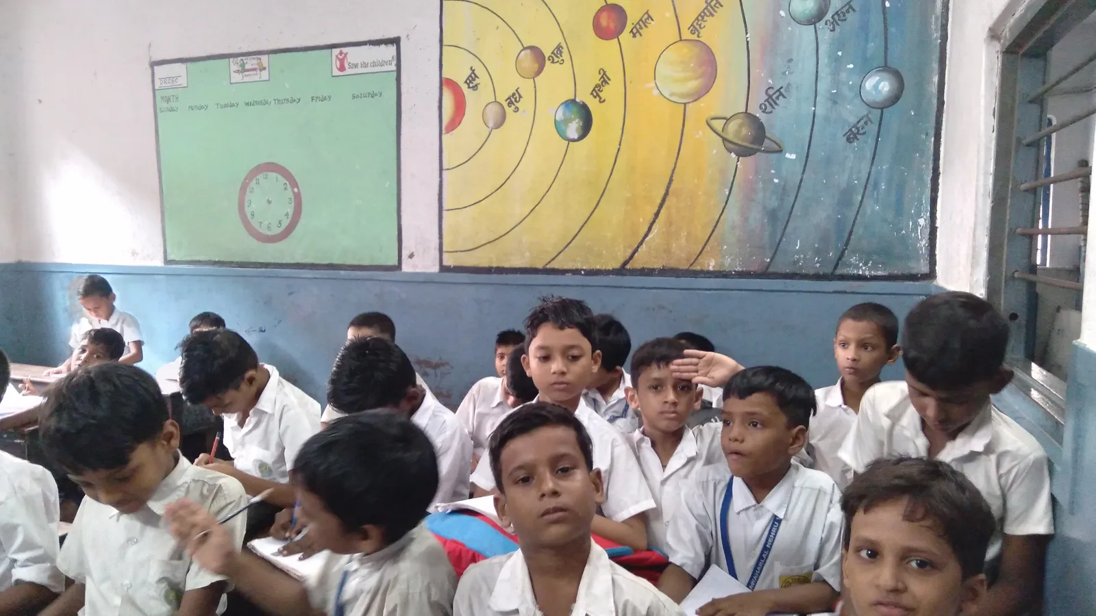 Crowded classroom of boys in white uniforms beneath a painted solar system mural
