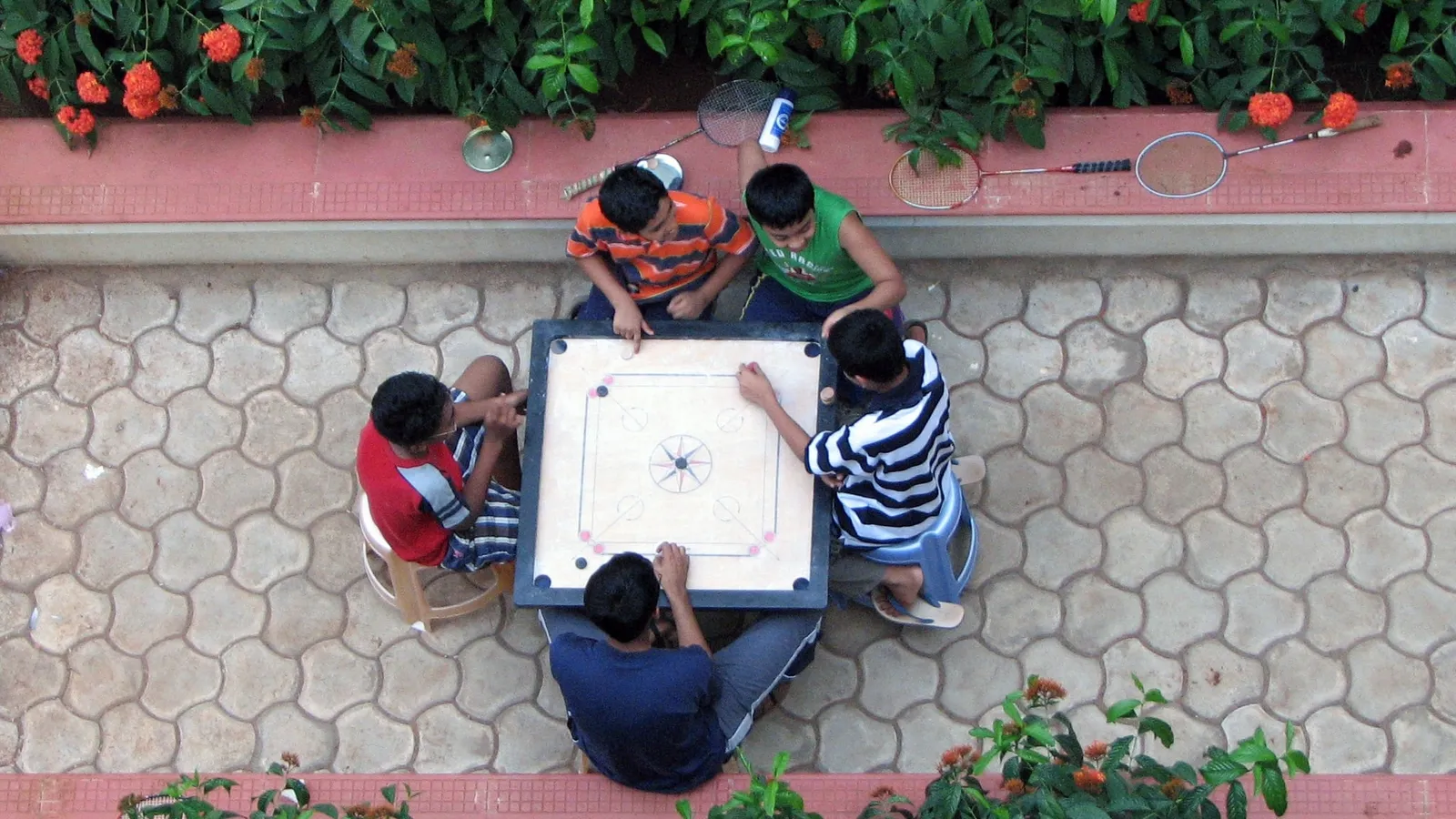 Five boys playing carrom in a paved courtyard, seen from above