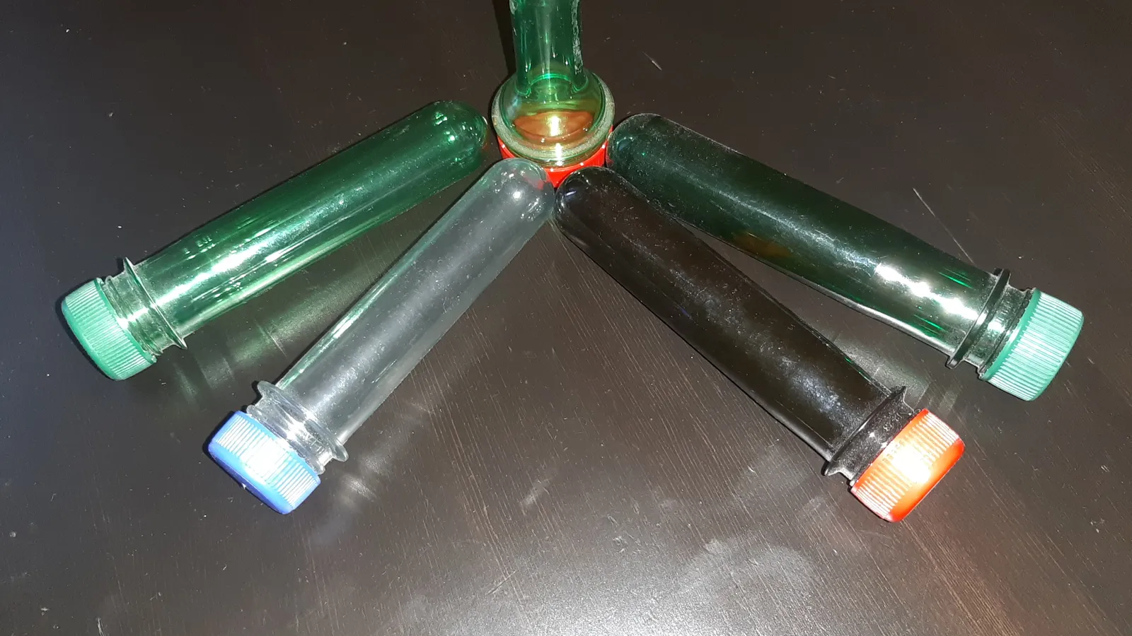 Five coloured PET bottle preforms with screw caps on a dark table