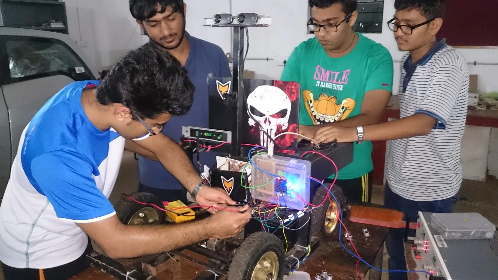 Four engineering students wiring a wheeled robot prototype in a college workshop
