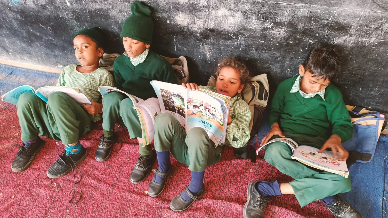 Four primary schoolchildren in green uniforms reading illustrated books against a blackboard