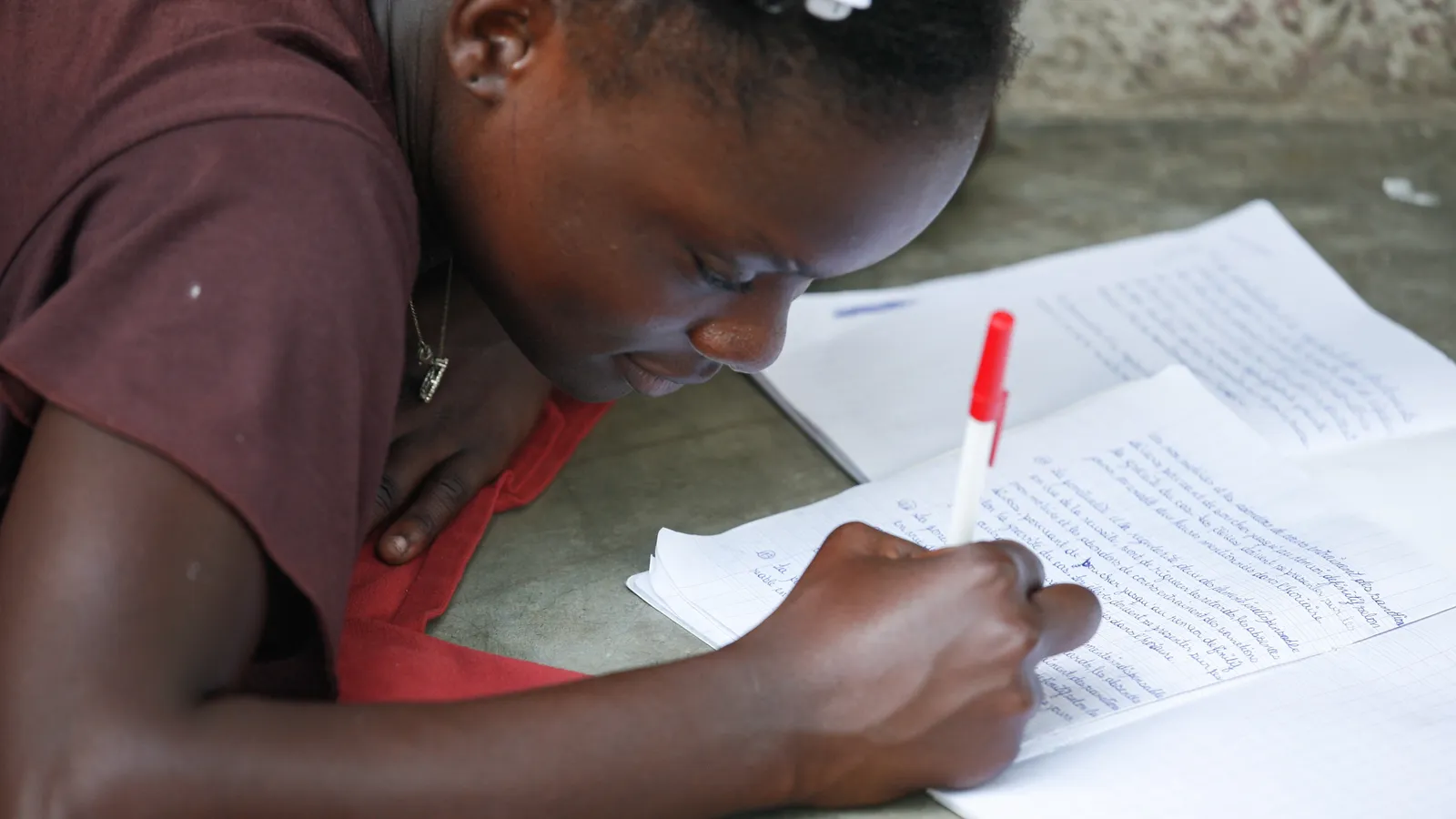 Girl bent over a notebook on a concrete floor, writing with a red pen