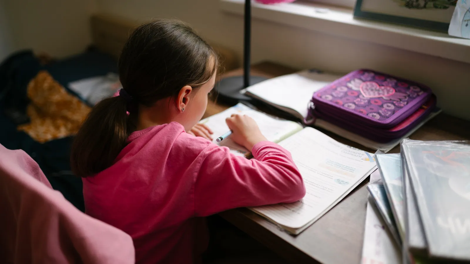 Girl in a pink top writing in a notebook at her study desk, seen from behind