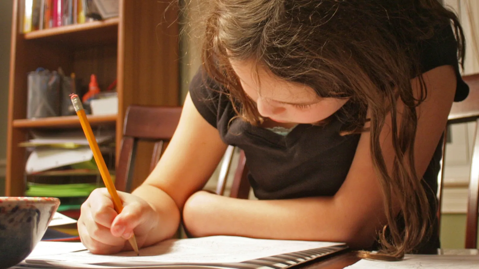 Girl leaning over a workbook at a wooden table, writing with a pencil